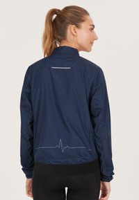 Marineblå windbreaker-jakke med strukturerede sidelapper, høj krave og et reflekterende hjertedesign på bagsiden; elastisk taljesyn.
