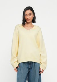 Απαλό κίτρινο πλεκτό πουλόβερ με V-λαιμόκοψη και υπερμεγέθη σιλουέτα. Διαθέτει μυτερό γιακά και ριμπ τελείωμα. Συνδυάζεται με άνετο μπλε τζιν.