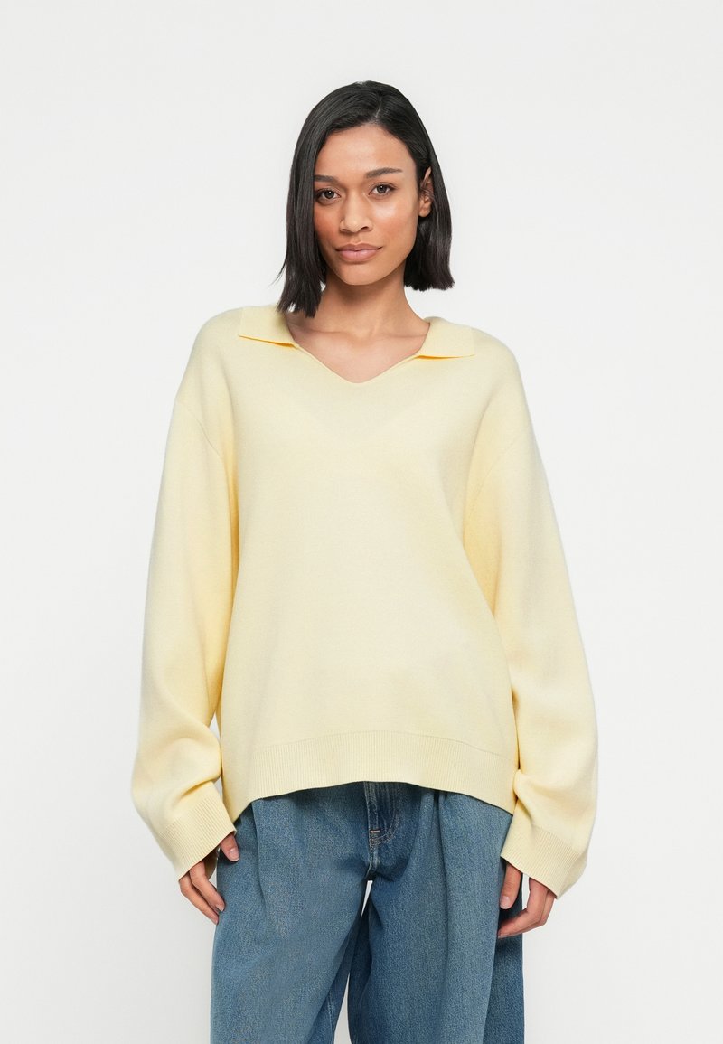 Απαλό κίτρινο πλεκτό πουλόβερ με V-λαιμόκοψη και υπερμεγέθη σιλουέτα. Διαθέτει μυτερό γιακά και ριμπ τελείωμα. Συνδυάζεται με άνετο μπλε τζιν.