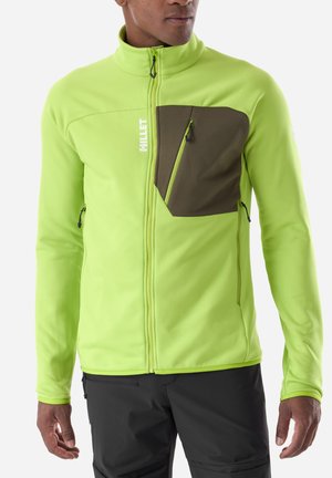Man wearing a bright green zip-up jacket with a dark green zippered chest pocket and black pants, standing with arms relaxed.