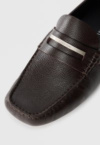 Brown leather loafer with textured surface, featuring a silver-toned logo plate and contrasting stitching along the edges.