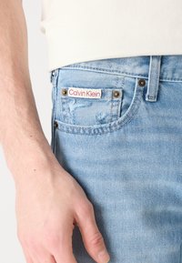 Ljusblå denimjeans med en sliten textur, försedda med en röd och vit Calvin Klein logotagsmärke, koppardetaljer och standard femficksdesign.