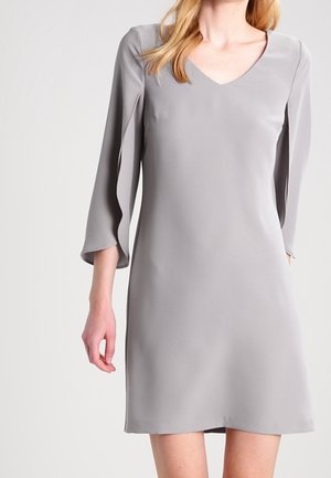 Femme portant une robe gris clair longueur genoux avec un col en V et des manches trois-quarts.