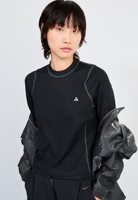 Black long-sleeve top with ribbed texture, round neckline, and contrasting stitching. Features small logo and layered with a shiny black jacket.
