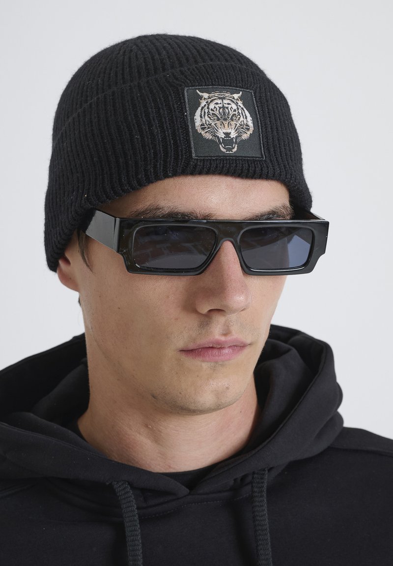 Zwarte gebreide beanie met een tijgergezicht patch; oversized zwarte zonnebril met hoekige frames; gedragen over een zwarte hoodie.