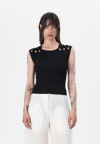 Black ribbed sleeveless top with a fitted design and gold button accents on the shoulders. Worn with white wide-leg trousers.