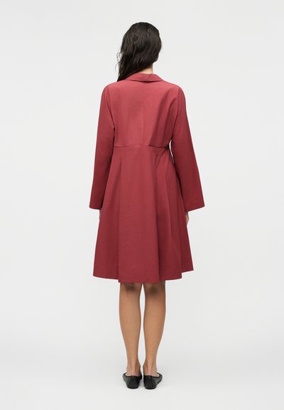 Γυναίκα που στέκεται με την πλάτη προς την κάμερα, φοράει παλτό μέχρι τα γόνατα, με μακριά μανίκια, σκούρο κόκκινο, και μαύρα παπούτσια slip-on σε απλό λευκό φόντο.