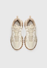 Zapatillas de malla beige con superposiciones texturizadas, que cuentan con suelas y cordones de goma marrón claro intricadamente tejidos con hilos reflectantes.