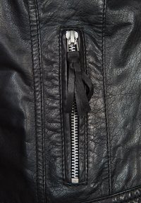 Detail černé kožené bundy se stříbrným zipem s texturovaným povrchem a koženým taháčkem, zdůrazňující šití a kvalitu materiálu.