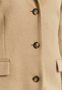 Beige wool coat featuring textured fabric, four dark buttons, and front pockets. Simple design with a classic fit and tailored lapels.