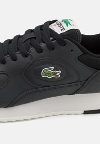 Baskets en cuir noir avec bout rond, lacets noirs, semelle blanche et petit logo crocodile vert. Dessus texturé avec des détails cousus.