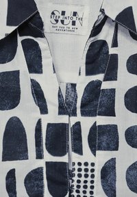 Tissu blanc avec un motif géométrique marine, comprenant des formes arrondies et ponctuées. Col classique avec des détails cousus au niveau de l'encolure.