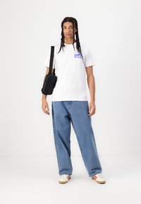 Hvit bomull t-skjorte med lilla grafisk tekst, sammen med løse lyseblå denimjeans. En liten svart skulderveske bæres på skrå.