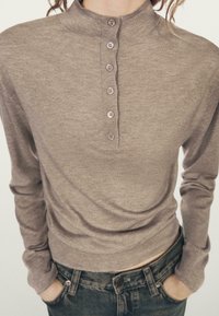 Brązowa bluzka z długim rękawem, wysokim kołnierzem i sześcioma guzikami. Miękka w dotyku, dopasowany fason. Noszona z ciemnymi dżinsami.