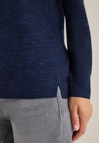 Dunkelblauer Strickpullover mit strukturiertem Muster, mit seitlichem Schlitz. Kombiniert mit hellgrauen Jeans. Nahaufnahme des Stoffs und der Hand.