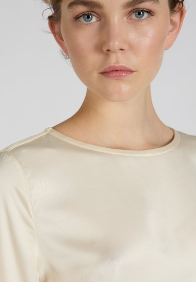 Helles cremefarbendes Satinoberteil mit glatter Textur, rundem Ausschnitt und kurzen Ärmeln. Verfügt über einen subtilen Glanz und ein minimalistisches Design.