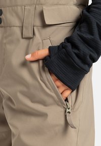 Pantalons de ski beiges avec une finition texturée et une taille ajustable. Comprend une poche zippée sur le côté et un haut à manches longues noir visible au niveau du poignet.
