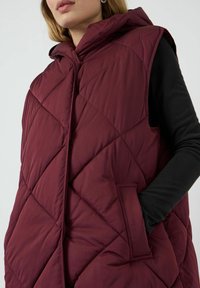 Burgunderfärgad pufferväst med huva, med en quiltad design och sidofickor. Tyget ser slätt och isolerat ut för värme.