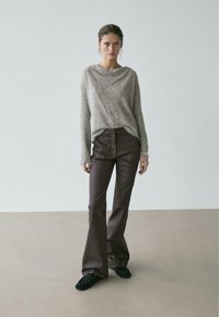 Kvinde står mod en ensfarvet baggrund iført en grå strikket sweater, mørkebrune læderbukser med vidde forneden og sorte mokkasinsko.