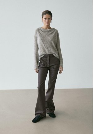 Donna in piedi davanti a uno sfondo semplice che indossa un maglione grigio a maglia, pantaloni a zampa di elefante in pelle marrone scuro e mocassini neri.