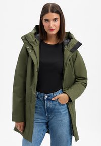 Olijfgroene waterdichte jas met een afneembare capuchon, ritssluiting en zichtbare zakken. Gecombineerd met een zwarte top en lichtblauwe jeans.