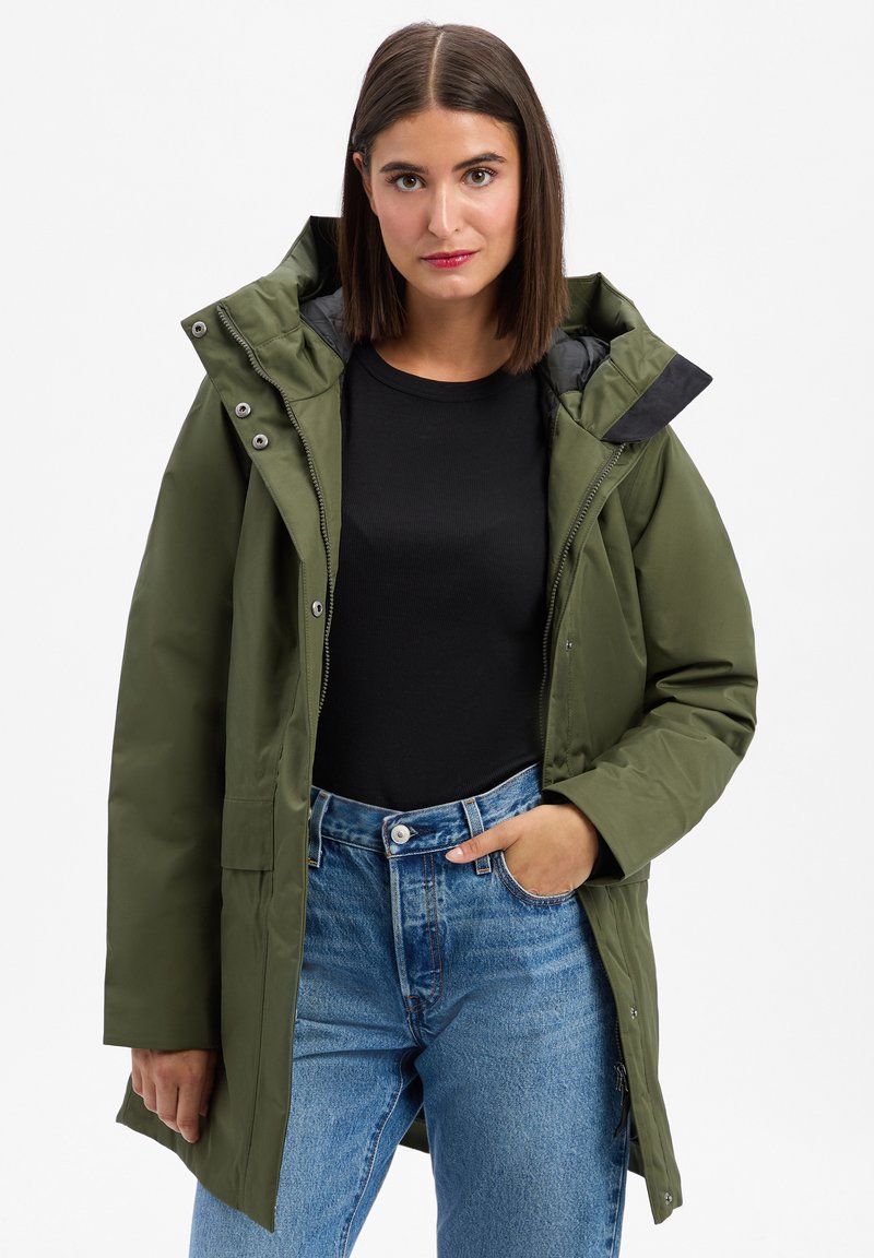 Olijfgroene waterdichte jas met een afneembare capuchon, ritssluiting en zichtbare zakken. Gecombineerd met een zwarte top en lichtblauwe jeans.