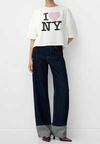 Camiseta blanca corta con "I ❤️ NY" en letras negras y rojas. Pantalones vaqueros de pierna ancha de un azul oscuro con puños grises y detalles de costura visibles. Tacones marrones.