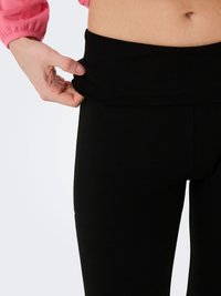 Person trägt schwarze Leggings und zieht den Bund nach außen, um die Dehnbarkeit zu zeigen; sichtbarer Bauchbereich und Hand mit rosa Ärmelsaum eines langärmeligen Oberteils.