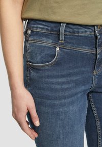 Jeans en denim bleu foncé avec une coupe slim, dotés d'une poche avant, de détails cousus, et d'une taille visible avec un bouton et une fermeture à boucle.