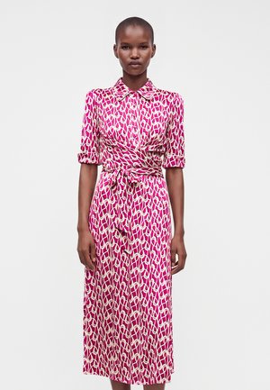 Femme portant une robe midi rose et blanche à motifs avec col, manches courtes, boutons et ceinture à la taille, debout devant un fond uni.