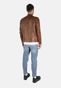 Giacca di pelle marrone con un design aderente, colletto alto e dettagli di cucitura sottili, abbinata a jeans in denim azzurro chiaro e sneakers nere.