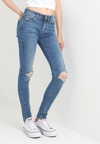 Jeans skinny de mezclilla azul con desgarros en las rodillas, con una cintura de altura media y un diseño ajustado. Combinados con zapatillas de lona blancas.