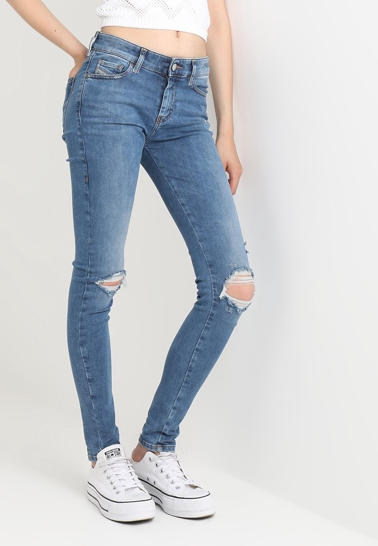 Jeans skinny de mezclilla azul con desgarros en las rodillas, con una cintura de altura media y un diseño ajustado. Combinados con zapatillas de lona blancas.