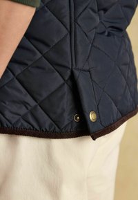 Námornícky prešívaný vest s hnedým kordurovým lemovaním, ktorý má kovové cvočky na dolnom leme. Textúra vyzerá hladko a vypchato, poskytuje izoláciu.