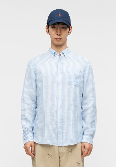 Camicia a righe azzurro chiaro con bottoni e maniche lunghe, caratterizzata da una tasca sul petto a sinistra e un piccolo logo, abbinata a un berretto da baseball blu scuro.