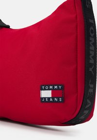  Bolsa de hombro de tela roja con una correa negra, un parche de logo rectangular en colores azul, blanco y rojo, y una superficie texturizada.