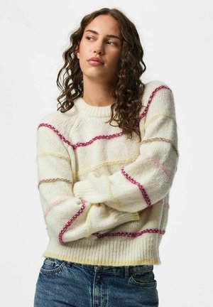 Femme aux cheveux bouclés portant un pull crème avec des rayures horizontales multicolores et un jean bleu, bras croisés, regardant sur le côté.