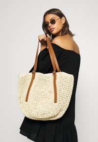 Borsa a spalla in paglia intrecciata di colore beige chiaro, con manici in pelle marrone e fibbie regolabili. Presenta un design in tessuto aperto e testurizzato.
