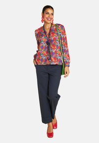 Smiling woman in a colorful floral blouse and navy pants, wearing red heart-shaped earrings and red heels, holding a green bag.