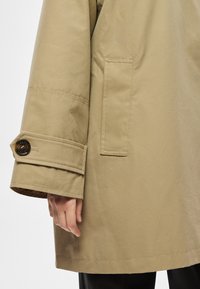 Bézs színű, laza szabású felsőruházat, gombos mandzsettával és vízszintes zsebbel. Sima, tartós anyag finom textúrával.