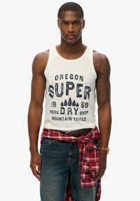 Witte tanktop met marineblauwe print van "Oregon SUPER DRY" en bomen. Draagt een blauwe spijkerbroek met een rood geruit shirt dat om de taille is gebonden.