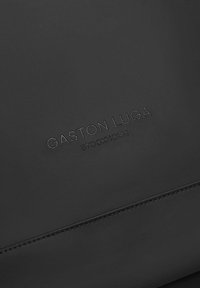 Black textured leather surface featuring embossed "GASTON LUGA" and "STOCKHOLM" branding. Subtle stitching detail adds dimension.