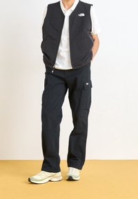 Chaleco negro sin mangas con cremallera, logo en el pecho, combinado con pantalones cargo negros con múltiples bolsillos, y zapatillas deportivas verde claro.