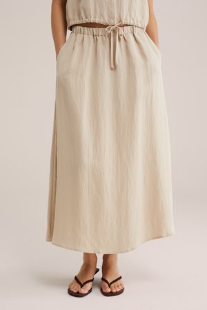 Beige midi-rok met hoge taille, trekkoord en zijzakken, gedragen met donkerbruine teenslippers op een neutrale achtergrond.