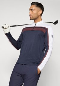 Námořnické golfové tričko s dlouhým rukávem s vínovými akcenty, zipovým límcem a bílými rukávy. Stylizováno se šedými kalhotami a golfovou holí.
