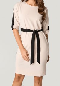 Beżowa sukienka z krótkimi czarnymi rękawami i dopasowanym czarnym paskiem związanym w talii. Gładki materiał o luźnym kroju i bocznymi kieszeniami.