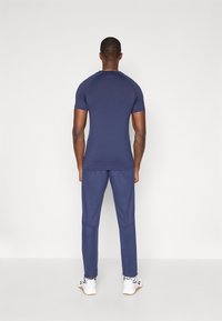 Ναυτικό αθλητικό μπλουζάκι με κοντά μανίκια και ταιριαστά φόρμα, κατασκευασμένο από μαλακό ύφασμα. Φορεμένο με άσπρα αθλητικά παπούτσια, απεικονίζεται από πίσω.