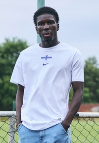 Weißes Baumwoll-T-Shirt mit blauem "ENG"-Logo und kleinem Nike-Swoosh. Das Model trägt hellblaue Jeansshorts und steht an einem Zaun.