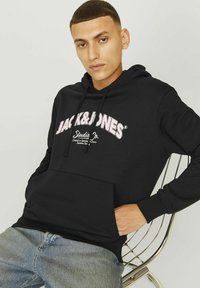 Crna dukserica s kapuljačom i prednjim džepom, s bijelim i ružičastim natpisom "JACK & JONES" i "Studio Co." u zakrivljenom dizajnu. Pamuk, ležeran kroj.
