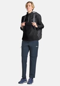 Pullover in pile nero con mezza zip, abbinato a pantaloni softshell blu. L'outfit è completato da uno zaino grigio e scarpe da trekking blu.